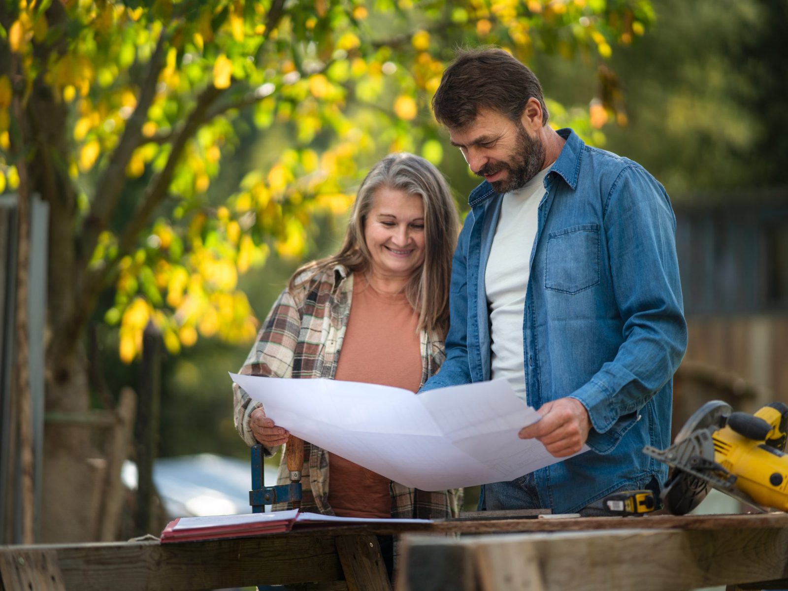 A happy mature couple with architectural blueprints of their future house, standing outdoors.