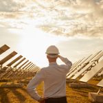 Back view of anonymous man in hardhat touching head and looking at modern photovoltaic panels and cloudy sundown sky during work on solar power station